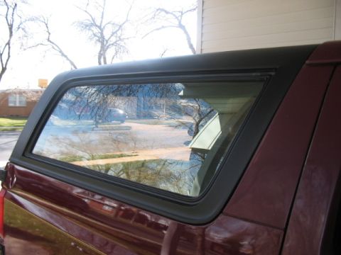 1991 Ford Bronco - The side window of a pickup truck shows reflections of trees and a house. A camper shell is attached to the truck bed.