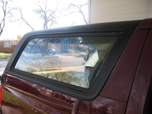 Side view of a maroon pickup truck with a black camper shell. A car is visible in the reflection of the window