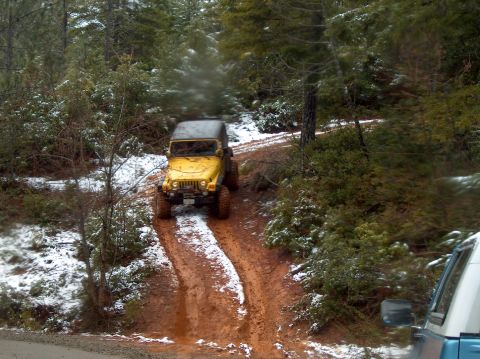 1993 Ford Bronco - A yellow Jeep navigates a muddy and snowy trail through a wooded area. Another vehicle is partially visible on the right side of the frame.