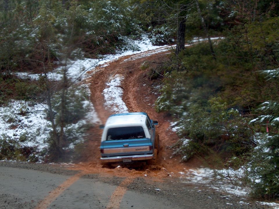 Blue and white SUV driving up muddy red-dirt trail through snowy woods