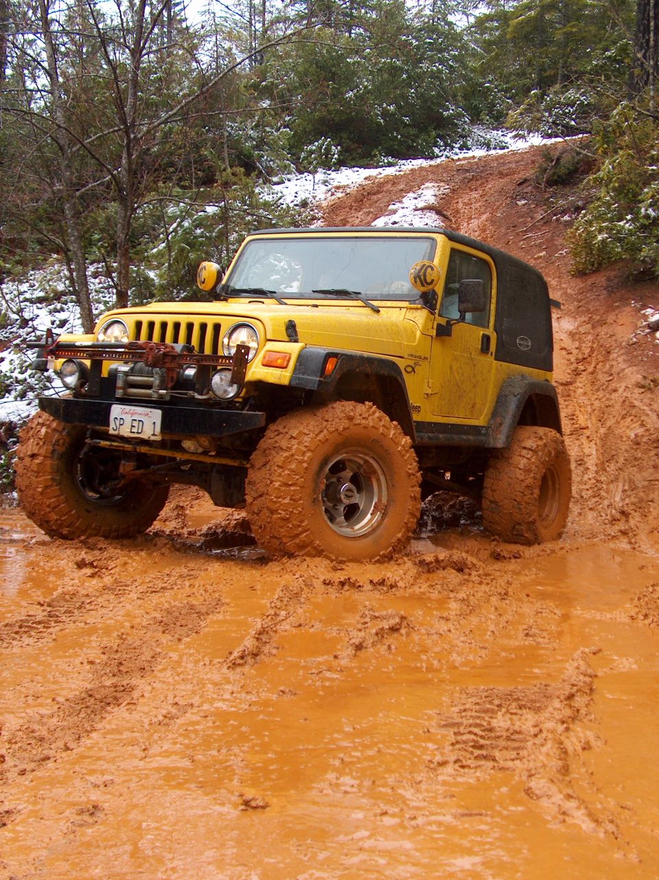 Yellow Jeep Wrangler driving through a muddy trail with snow on the trees