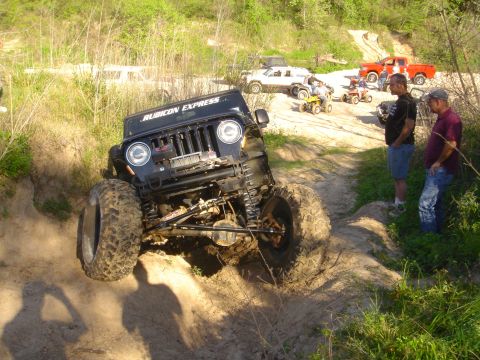 2002 Jeep Wrangler - A group of off-road vehicles, including ATVs and trucks, gather on a dirt path while a modified Jeep Wrangler tackles an incline.