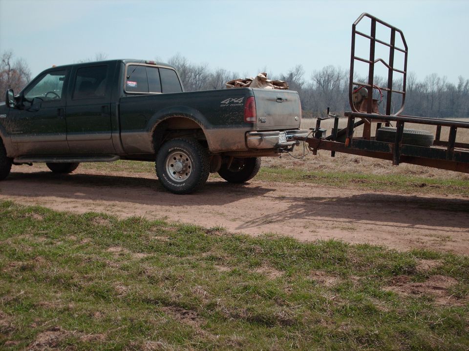 Muddy dark green Ford 4x4 crew cab pickup towing a flatbed trailer on a dirt path in a field