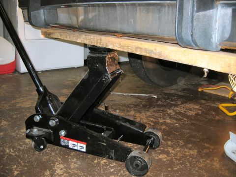 1985 Ford LTD Crown Victoria - A car's side panel and wheel are elevated by a floor jack and a wooden plank.