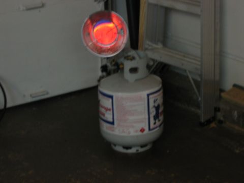 1985 Ford LTD Crown Victoria - A propane tank sits on the floor with a heater element glowing red above it, near a ladder in a garage.