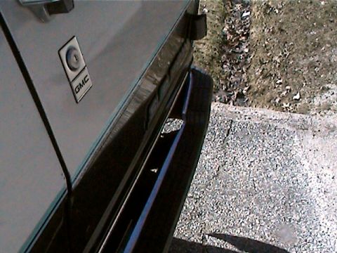1977 Ford F-150 - A close-up view of a GMC emblem on the side of a truck, with gravel and dry grass in the background.