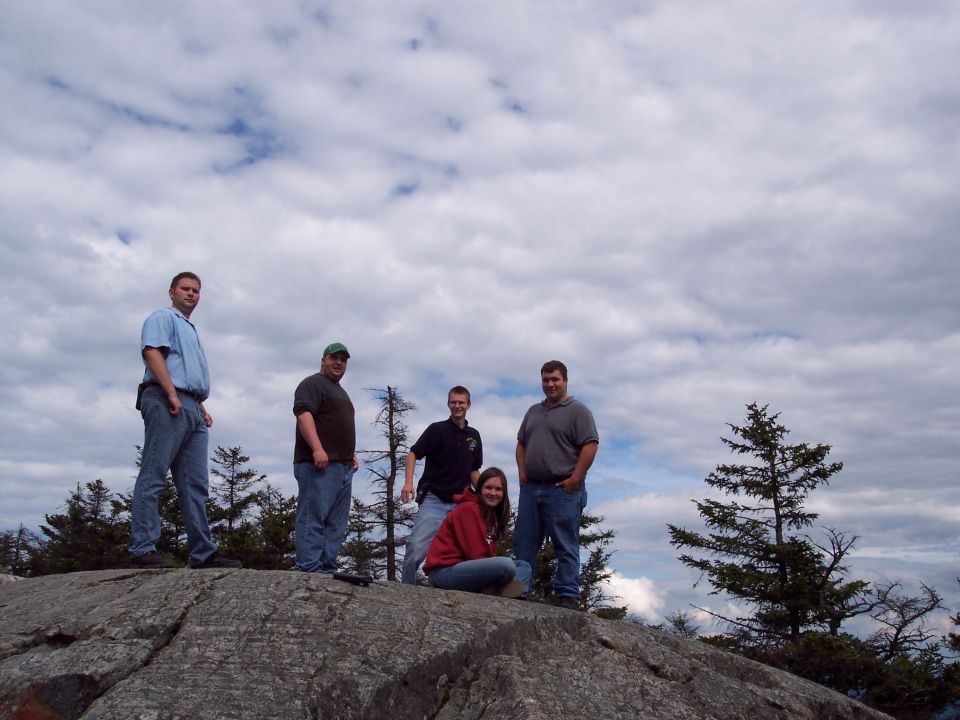 Group of five people standing and sitting on a large rock formation with trees and a cloudy sky in the background