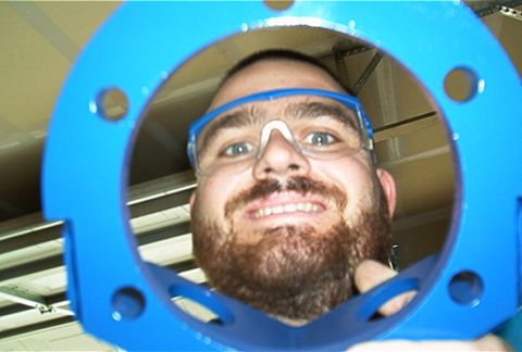 1986 Ford Bronco - Man smiles through a blue metal part with bolt holes, wearing safety glasses.