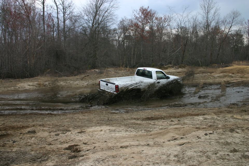 White regular-cab pickup splashing through a mud hole in a sandy off-road area with bare trees behind