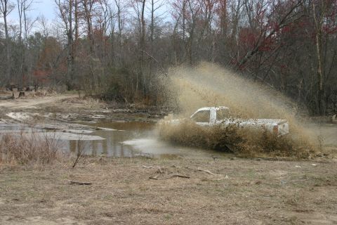 1989 Ford Bronco - A white pickup truck navigates a muddy obstacle, creating a large spray.