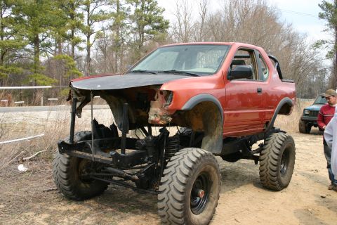 1989 Ford Bronco - A heavily modified orange off-road vehicle sits in a dirt lot with another vehicle in the background.