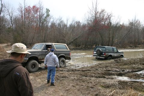 1989 Ford Bronco - Two Ford Broncos navigate a muddy off-road course, with one vehicle partially submerged in water.