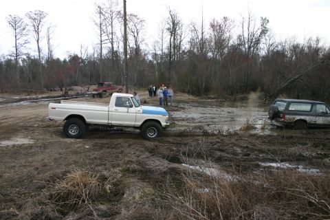 1989 Ford Bronco - Two trucks are in a muddy, wooded area. One is driving on a dirt path, while the other is stuck in the mud.