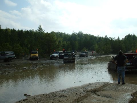 1991 Ford Bronco - Multiple off-road vehicles traverse a challenging muddy and watery course under a cloudy sky.