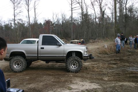 1989 Ford Bronco - Silver pickup truck with lifted suspension and large off-road tires sits in a muddy area with trees and people around.