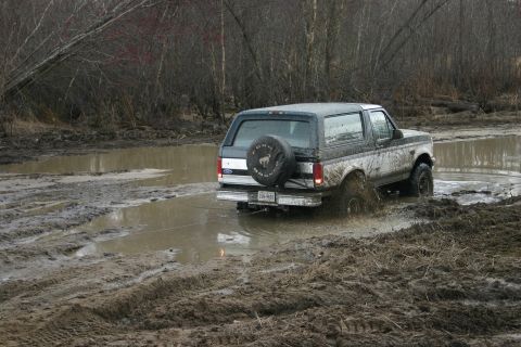 1989 Ford Bronco - This Ford Bronco is navigating a challenging muddy terrain, kicking up water and dirt.