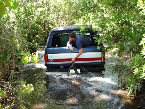 1989 Ford Bronco - A man attempts to free a blue and white Ford Bronco stuck in a watery, overgrown area.