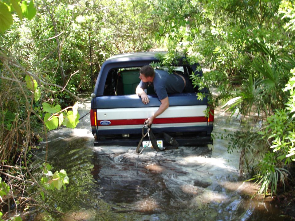 Blue and white Ford Bronco with red stripe stuck in water, man leaning out rear window