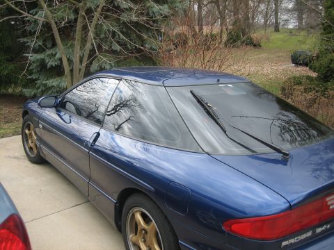 1995 Ford Probe - A blue coupe with gold rims is parked on a concrete driveway, with trees and a grassy hill in the background.