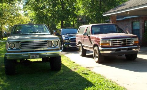 1996 Ford Bronco - Three trucks lined up in a residential yard: a lifted tan square-body Dodge pickup on mud tires wearing a Texas plate reading 98B-DCZ at le…