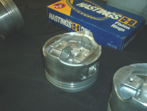 1994 Mazda MX-3 - Engine pistons and a box of Hastings piston rings are displayed on a dark surface.