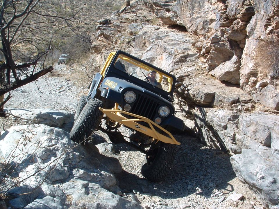 Jeep CJ-7 with yellow roll cage and bumper climbing over rocks on a trail. Another SUV is visible in the distance