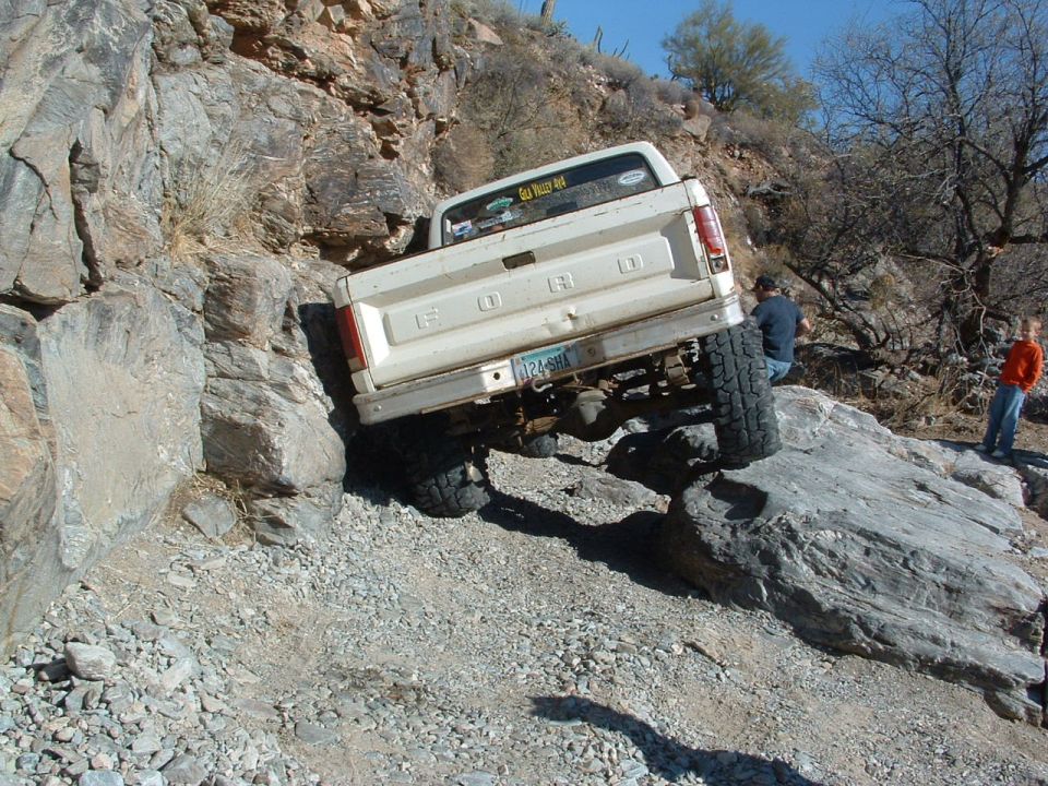 White Ford pickup truck with a lifted suspension is driving over rocks. The truck has a dent on the tailgate