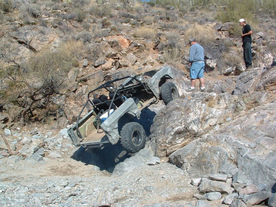 Off-road buggy with roll cage navigating a steep, rocky hill with two people watching