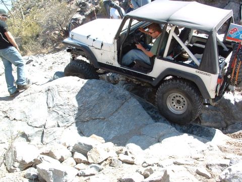 1981 Ford F-150 - A white Jeep Wrangler maneuvers over large rocks on an off-road trail, with a Domino's Pizza logo visible on the spare tire cover.