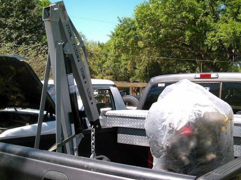 1986 Ford Bronco - An engine hoist is positioned in a truck bed, with a large, covered item nearby and another vehicle in the background.