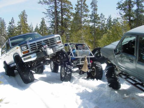 1986 Ford Bronco - Multiple modified off-road vehicles navigate a snowy terrain with trees in the background.
