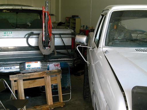 Two pickup trucks, one white and one silver, parked in a garage. A Hi-Lift jack is mounted on the silver truck's tailgate