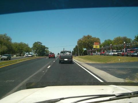 1986 Ford Bronco - A pickup truck drives down a highway past a car dealership on a sunny day.