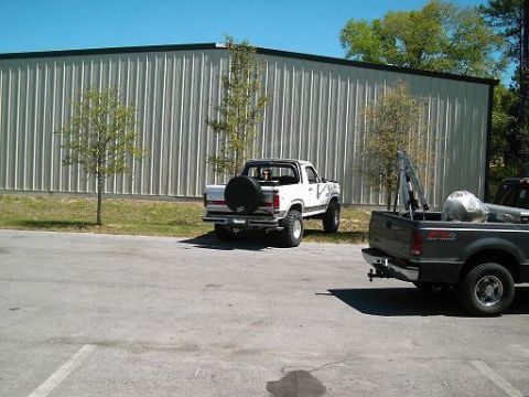 1986 Ford Bronco - A white pickup truck is parked outside a large metal building, with another truck in the foreground.