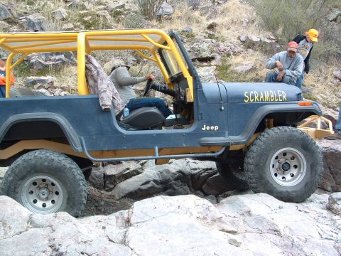 1981 Ford F-150 - A blue Jeep CJ-8 Scrambler, equipped with a yellow roll cage, is shown off-roading over a rocky landscape.