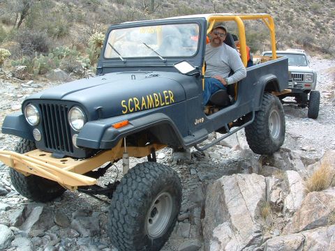 1981 Ford F-150 - A man drives a dark blue Jeep CJ-8 Scrambler off-road over rocky terrain, with another vehicle following behind.