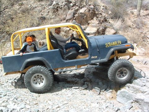 1981 Ford F-150 - A blue Jeep Scrambler, identified by its 'SCRAMBLER' badging, navigates a rocky trail with a family and dog inside.