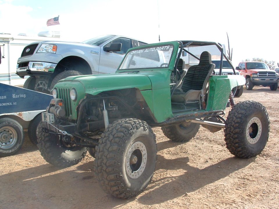 Green off-road jeep with large tires and roll cage, parked next to a silver Ford F-150