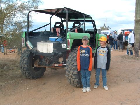 1981 Ford F-150 - Two boys stand next to a heavily modified off-road vehicle in a desert environment with other vehicles present.