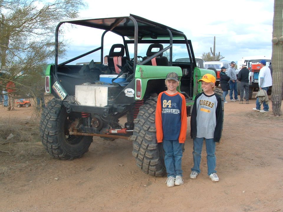 Modified off-road vehicle with roll cage, large tires, and two boys standing next to it. Other vehicles and people in background