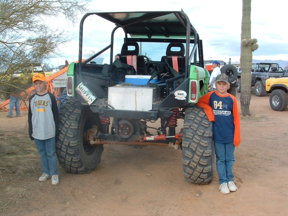 Green off-road vehicle with roll cage, large tires, and two children standing next to it. Visible text: MUFFIN, RWC, SUPERMOTORS.NET