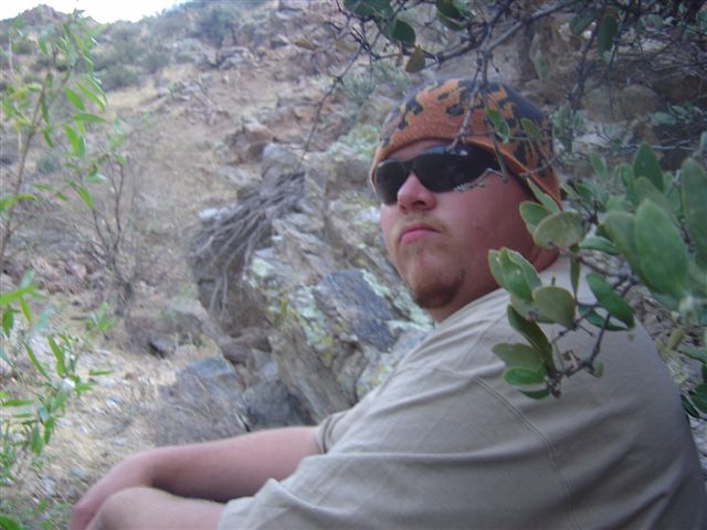Man wearing a beanie and sunglasses sits in a rocky, desert landscape with sparse vegetation