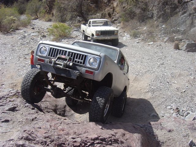 Silver International Scout off-roading, with a white pickup truck behind it on a rocky trail