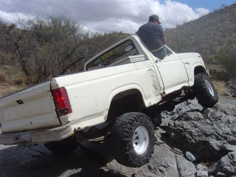 1981 Ford F-150 - A white pickup truck is shown off-roading on a rocky trail with a person sitting on the open driver's door.