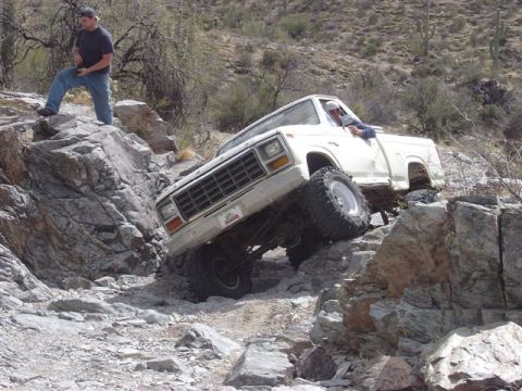 1981 Ford F-150 - A white 1980 Ford F-150 is shown tackling a challenging rocky off-road course with a person standing on a boulder nearby.