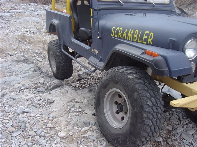 Dark blue Jeep CJ-8 Scrambler with yellow "SCRAMBLER" text on the hood, off-road tires, and yellow roll bar