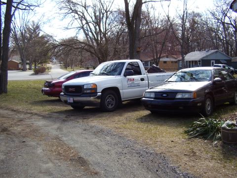 1997 Ford Taurus - A dark sedan is parked in a driveway next to a white pickup truck and a red car.