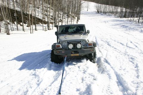 2003 Jeep TJ - A silver 2003 Jeep TJ is stuck in deep snow on a wooded trail, its front end facing the camera with a tow strap attached.
