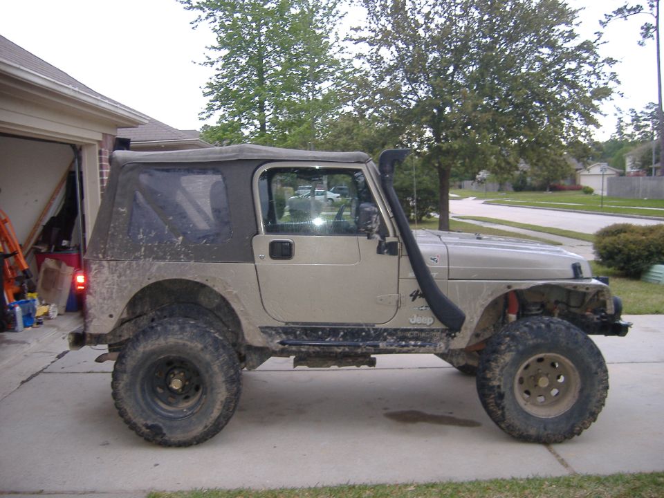 Side view of a tan Jeep Wrangler with a snorkel, soft top, and large tires parked in a driveway