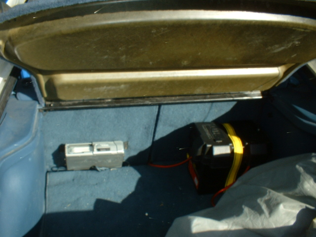 Car trunk interior showing a battery with yellow straps and a small silver device on the left